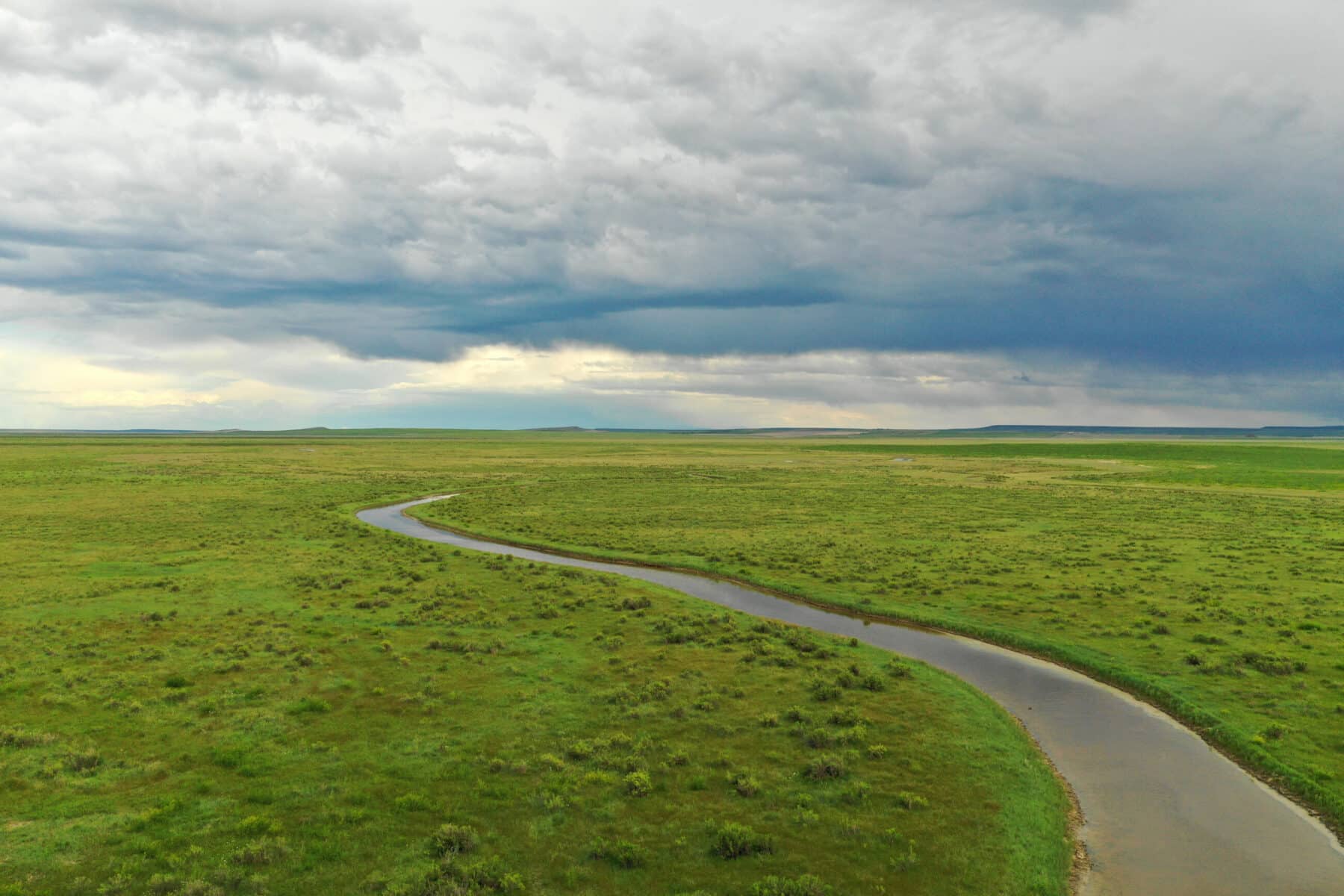 A winding river flows through a vast, green grassy plain under a cloudy sky, with dark storm clouds gathering on the horizon—a perfect setting for a picturesque ranch for sale.